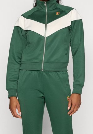 Personne portant un survêtement vert avec un chevron blanc sur la poitrine et un petit logo coloré sur la cuisse gauche et droite.