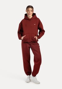 Bordeauxroter Kapuzenpullover und passende Jogginghose aus weichem Stoff. Der Hoodie verfügt über eine Vorderasche und ein kleines Logo. Sneakers vervollständigen das Outfit.
