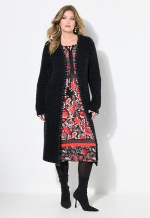 Cardigan long noir en peluche, robe florale avec des motifs rouges et blancs, accents noirs, et bottes noires pointues jusqu'aux genoux.