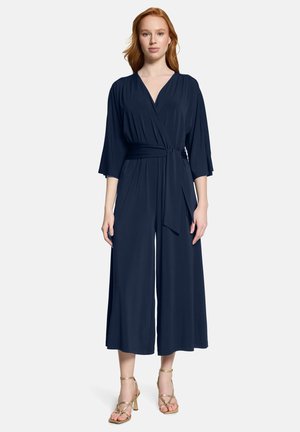 Kobieta o rudych włosach ubrana w granatowy kombinezon z kopertowym zapięciem, szerokimi nogawkami i rękawami do łokcia, w parze ze złotymi sandałami na paskach.