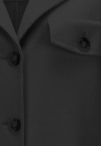 Schwarzer Wollmantel mit Reverskragen, zwei großen Knöpfen und einer einzelnen Brusttasche. Weiche Textur und strukturierte Optik.