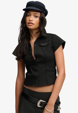 Ung kvinde med langt mørkt hår iført en sort crop top med striber, matchende nederdel, sort hat og sølv bæltespænde.