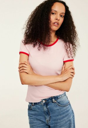 Młoda kobieta o kręconych włosach, ubrana w różowy t-shirt z czerwonym wykończeniem i niebieskie jeansy, stoi z założonymi rękami na tle jednolitego tła.
