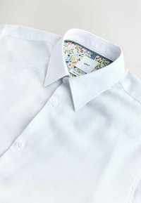 Biała koszula zapinana na guziki, z ostrym kołnierzykiem i kolorowym kwiatowym wzorem wewnątrz kołnierza, oznaczona jako "Next" i "100% bawełna".