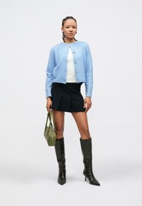 Vaaleansininen neulemallinen neuletakki kultaisilla napeilla, valkoinen paita, musta pliseerattu minihame, vihreä käsilaukku ja mustat polven korkeuteen ulottuvat korkokengät.