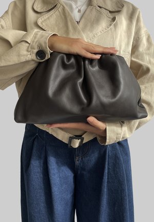 Borsa a clutcher in pelle nera con design ripiegato, tenuta in una mano. Presenta una texture liscia e bordi morbidi e arrotondati. Indossata con un giacca beige.