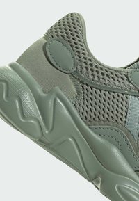 Green athletic shoe featuring mesh and smooth synthetic material, a textured sole with wave patterns, and a reinforced heel tab.