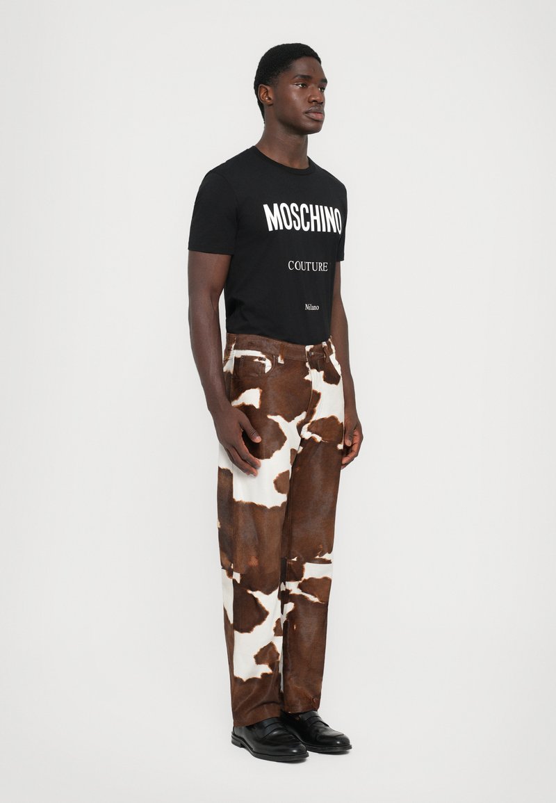 Schwarzes Baumwoll-T-Shirt mit weißem "Moschino Couture"-Text, kombiniert mit creme- und braunem Kuhflecken-Hosen in weitem Schnitt. Schwarze Lederschuhe vervollständigen den Look.