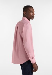 Camicia a maniche lunghe con bottoni in tessuto rosa chiaro dalle texture sottile. Presenta un colletto classico e polsini con bottoni. Indossata con jeans scuri.