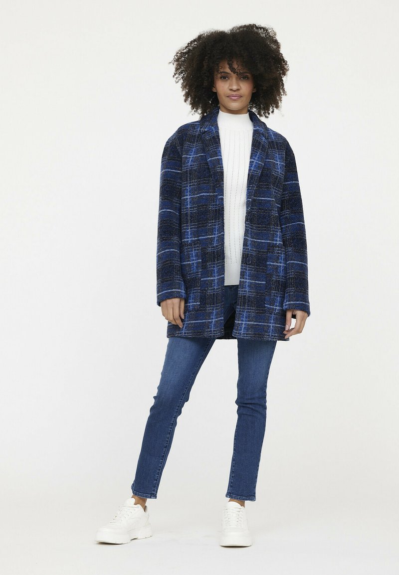 Blauw en zwart geruite jas met een relaxed fit, met lange mouwen, een sjawlkraag en knoopsluiting. Gemaakt over een witte trui en jeans.