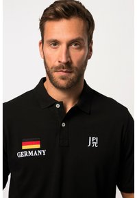 Černé polo tričko s nášivkou německé vlajky, vyšitým textem "NĚMECKO" a bílým logem. Dvoubottonový límec a krátké rukávy.