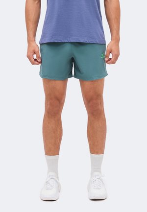 Άνδρας που φοράει τιρκουάζ σορτς Nike, λευκές κάλτσες, λευκά αθλητικά παπούτσια και μπλε πουκάμισο, στέκεται μπροστά σε μια απλή ανοιχτή επιφάνεια.