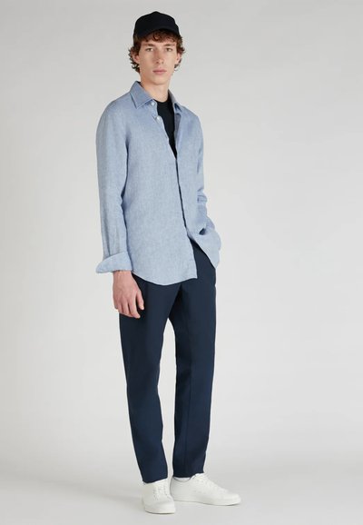 Camisa de botón azul claro, mangas largas arremangadas, combinada con pantalones navy y zapatillas blancas. Fondo neutro.