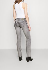 Donna che indossa jeans grigi slim, top senza maniche bianco e stivaletti neri alla caviglia, in piedi di spalle su uno sfondo chiaro e uniforme.