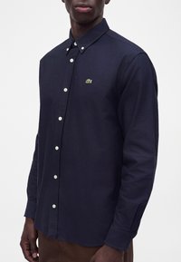 Homme portant une chemise bleu foncé boutonnée avec des boutons blancs et un petit logo crocodile vert sur la poitrine.