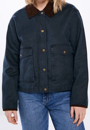 Chaqueta azul marino con cuello y puños de pana marrón, botones de presión delanteros, dos bolsillos con solapa, combinada con jeans azules.