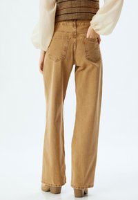 Jeans de tiro alto de color marrón claro con pierna recta, con bolsillos traseros y una textura ligeramente descolorida. Combinando con una blusa crema.