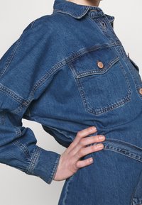 Denimoverall i mörkblått med krage, knäppta bröstfickor och långa ärmar. Har synliga sömmar och en avslappnad passform.