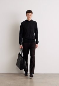 Schwarzer Strickpullover mit Raglanärmeln, kombiniert mit dunkelbraunen Hosen. Das Model trägt eine schwarze Tote-Tasche mit geprägtem Logo und steht vor einer schlichten Wand.