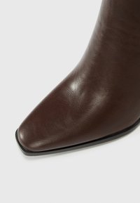 Bottine en cuir marron avec un bout pointu, texture lisse et design minimaliste. Elle présente un talon noir fin et aucun matériel visible.