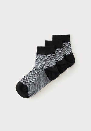 Trois paires de chaussettes noires et grises à cheville avec un motif en zigzag empilées sur un fond blanc.
