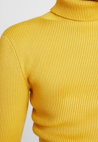 Gros plan d'un pull à col roulé jaune en maille côtelée montrant l'épaule, la manche et la partie supérieure de la poitrine.