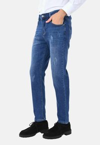 Jeans in denim blu con una vestibilità slim, caratterizzati da dettagli usurati e una texture sbiadita. Abbinati a scarpe in suede nere.