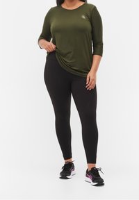 Maglietta verde oliva a maniche lunghe con scollo rotondo e drappeggio laterale, abbinata a leggings neri. La modella indossa scarpe da ginnastica nere e viola.
