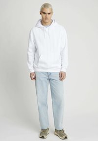 Haut à capuche blanc avec des rayures verticales, poche kangourou et cordon de serrage. Associé à un jean bleu clair et des baskets grises.