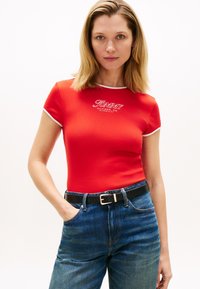 Ερυθρό εφαρμοστό κοντομάνικο μπλουζάκι με λευκή περιφέρεια, με το λογότυπο «TOMMY HILFIGER» μπροστά, σε συνδυασμό με ψηλόμεσο μπλε τζιν και μαύρη ζώνη.