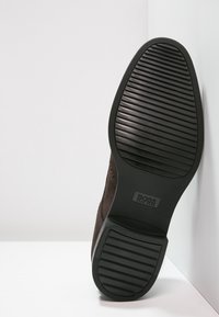 Chaussure en daim marron avec une semelle en caoutchouc texturée, présentant un dessous à motif pour une meilleure traction et un logo embossé sur la zone du talon.