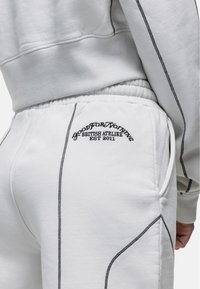 Witte sweatpants met een elastische tailleband, zwarte stikdetails en geborduurde branding op de achterzak. Zachte textuur.