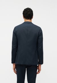 Άνδρας με κοντά σγουρά μαλλιά, ντυμένος με σκούρο ναυτικό μπλε σακάκι και αντίστοιχο παντελόνι, στέκεται μπροστά σε απλό λευκό φόντο.