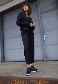 Kobieta z kręconymi włosami, ubrana w czarną koszulę, czarne spodnie i czarne trampki, stoi na betonowej platformie przed szarymi metalowymi drzwiami.