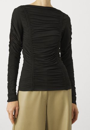 Personne portant un haut noir froncé à manches longues et un pantalon beige à jambes larges, visible du cou aux hanches sur un fond uni.