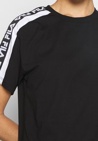 Svart t-shirt i bomull, med vita sidstreck och "FILA"-logotyp. Rundad halsringning och korta ärmar med en jämn textur.