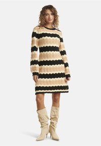 Eine Frau trägt ein knielanges, gewelltes, gestreiftes Kleid in Beige, Creme und Schwarz mit langen Ärmeln und beigen, lockeren, hohen Stiefeln.