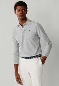 Muž oblečený v svetlosivej polo košeli s dlhým rukávom a malým logom, bielych nohaviciach a námorníckom prepletanom opasku, stojaci s jednou rukou v vrecku.