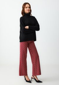 Maglione nero a collo alto, vestibilità ampia con texture a coste. Pantaloni a gamba larga rossi con motivi e accenti color crema e bordeaux. Decolleté neri a punta.