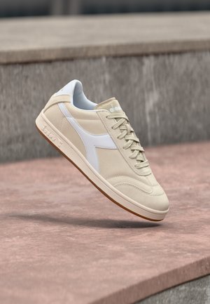 Béžová sportovní teniska s bílými doplňky, materiál z semiše a kůže, plochá gumová podrážka a symetrický šněrovací design. Jednoduchý a funkční tvar.