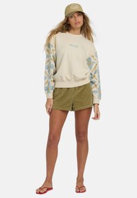 Sudadera crema con mangas estampadas en azul y dorado, combinada con pantalones cortos de pana verde oliva y chanclas rojas. Gorra beige con texto.
