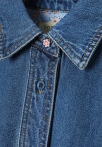 Col col en jean avec doublure en tissu floral et un petit bouton rose en forme de fleur près de la boutonnière supérieure.