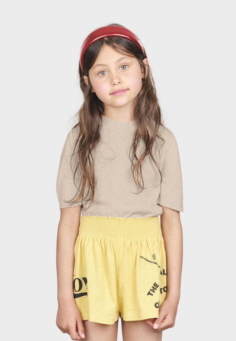 Suéter beige de manga corta, combinado con pantalones cortos amarillos de cintura elástica con gráficos de texto en negro. Cabello largo castaño con una diadema roja.