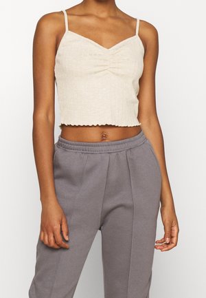Débardeur court côtelé beige clair avec fines bretelles, présentant un centre froncé et un ourlet ondulé, associé à un pantalon de survêtement gris.