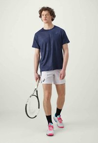 Camiseta deportiva con patrón navy, pantalones cortos blancos, calcetines negros, zapatillas de tenis coloridas. Sosteniendo una raqueta de tenis con un agarre blanco.