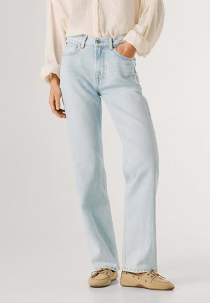 Mujer con vaqueros rectos azul claro, mocasines beige y una camisa crema de manga larga abotonada, con una mano en el bolsillo.