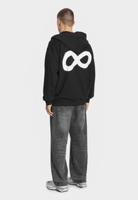 Czarna bluza z kapturem z dużym białym symbolem nieskończoności na plecach. W zestawie z szarymi szerokimi jeansami i jasnoszarymi sneakersami.