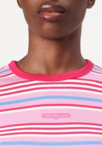 Striped t-shirt in pink, white, and blue. Features a round, pink ribbed collar and a "tommy jeans" logo in the center. Smooth fabric texture.