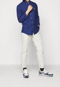 Mies, joka pukeutunut tummansiniseen, nappilistallinen pitkähihainen paitaan, luonnonvalkoisiin käärityihin housuihin, mustiin sukkiin ja valkoisiin lenkkareihin, joissa on sinisiä ja oransseja korostuksia.