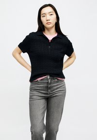 Young woman with long black hair wearing a black short-sleeve knit polo, pink shirt underneath, and grey high-waisted jeans, hands on hips.